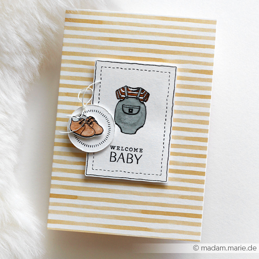 Grußkarte von Alexandra Renke mit beigen Streifen wie Designpapier Streifen senfgelb, mit Baby-Overall, Text "Welcome Baby" und einem Anhänger mit Babyschuhen - ideal für Papierprojekte oder Buchbinderei-Geschenke.
