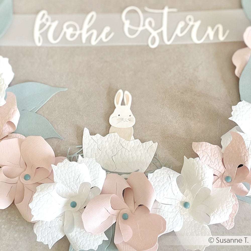 Papierkranz mit pastellrosa und weißen Blumen, einem Häschen und dem Text "Frohe Ostern" darüber, hergestellt aus FSC-zertifiziertem Karton für einen umweltfreundlichen festlichen Akzent.