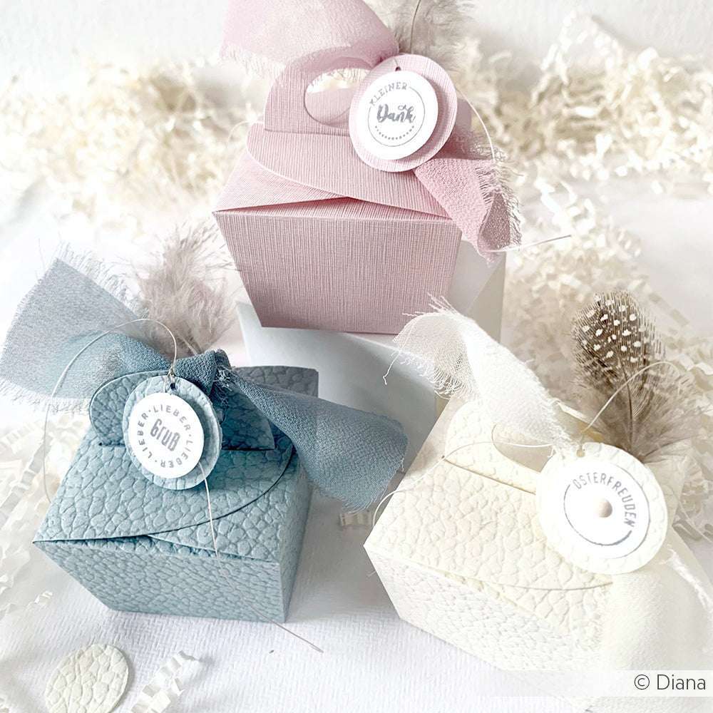 Drei pastellfarbene Geschenkboxen mit Bändern und Anhängern, umgeben von weißen Dekoschnipseln aus FSC-zertifiziertem 3D-geprägtem Papier von ☆, bringen Eleganz und nachhaltigen Stil in Ihre Geschenke.