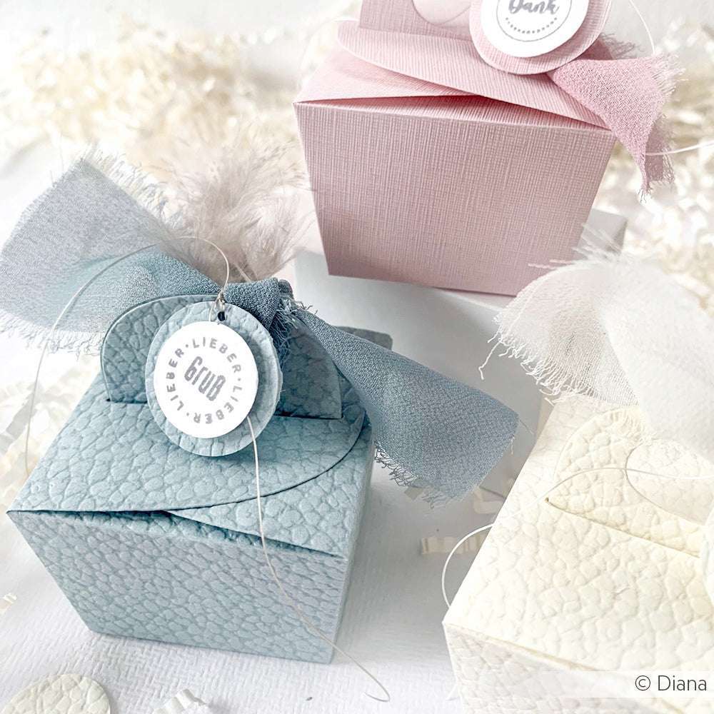 Drei strukturierte Geschenkboxen von ☆ in Pastellblau, Rosa und Creme, hergestellt aus FSC-zertifiziertem 3D-geprägtem Papier, jeweils verziert mit einem Band und einem runden Anhänger.
