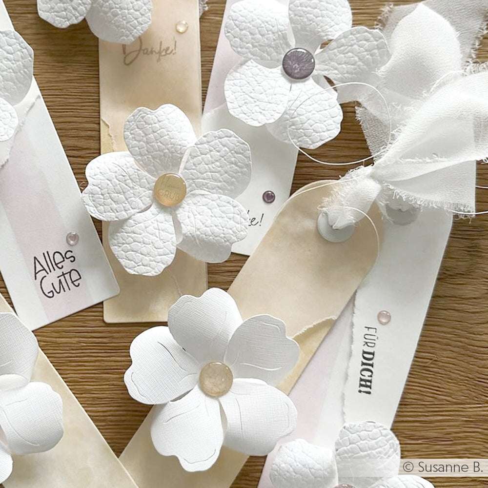 Die handgefertigten weißen Papierblumen aus FSC-zertifiziertem Karton schmücken Geschenkanhänger mit deutschen Botschaften und sind wunderschön auf einer Holzunterlage arrangiert.