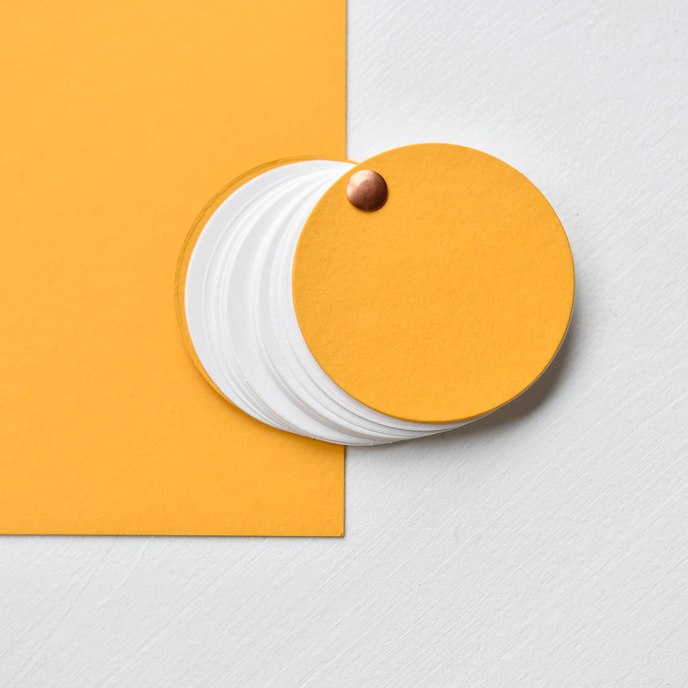 Auf einer weißen Fläche liegt ein Stapel gelber und weißer kreisförmiger Papierbahnen aus Alexandra Renke Uni Cardstock und Spezialpapier 700g/m², die von einer Messingklammer gehalten werden.