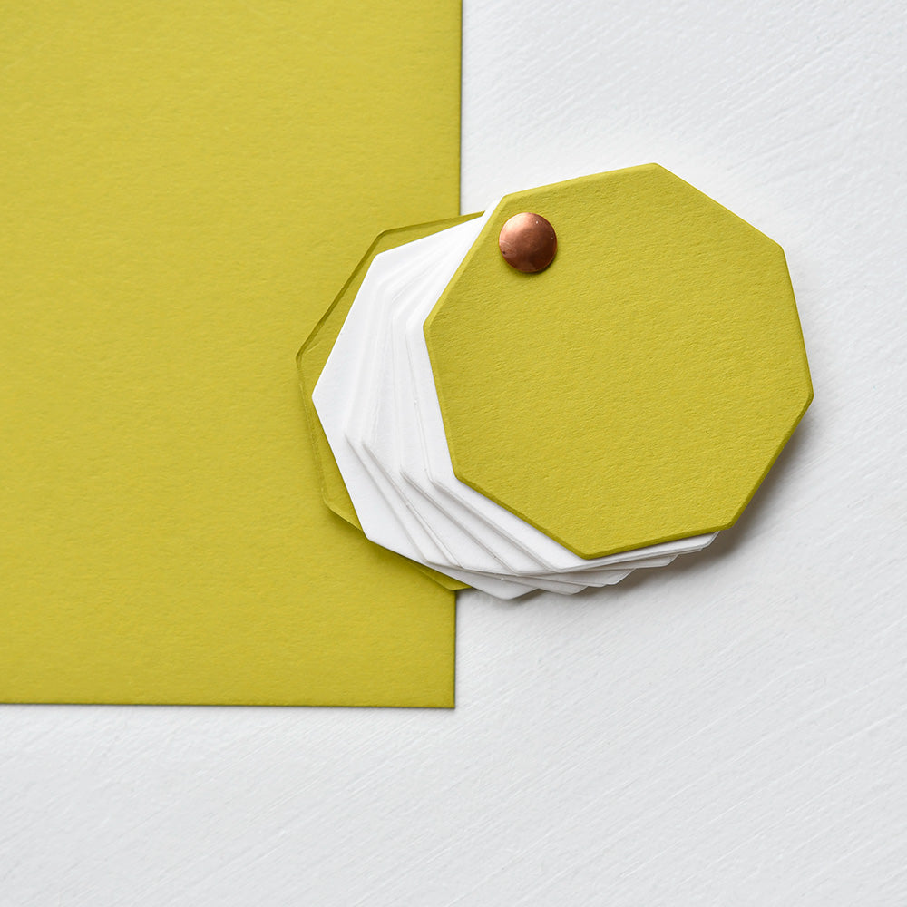 Auf einer weißen Fläche liegt ein Stapel gelber und weißer achteckiger Kartonbögen (700 g/m² Spezialpapier) von Alexandra Renke, die mit einer Messingklammer befestigt sind.