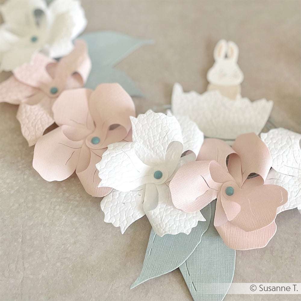 Pastellrosa und weiße Papierblumen mit blauer Mitte und grünen Blättern, hergestellt aus FSC-zertifiziertem Papier 120g/m² von ☆, arrangiert auf einer weichen beigen Oberfläche.