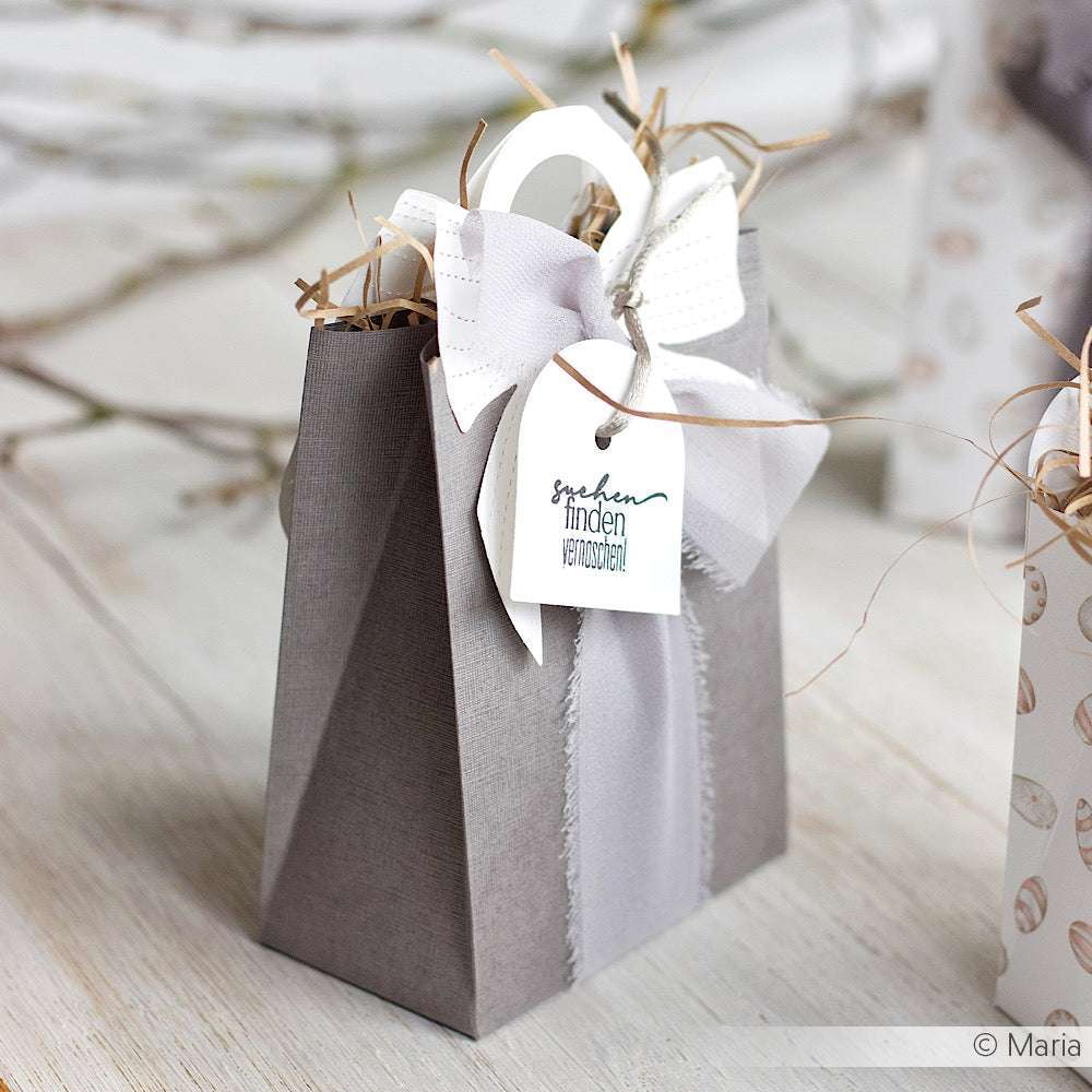 Graue Geschenktüte mit weißer Schleife, hergestellt aus FSC-zertifiziertem Papier 120g/m², gefüllt mit Stroh und mit einem Anhänger versehen; ausgestellt auf einer hellen Holzfläche.