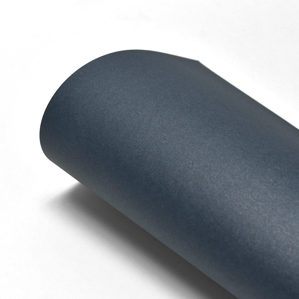 Ein aufgerolltes Blatt dunkelblaues ☆ 310g/m² Papier mit Schimmereffekt liegt auf einer weißen Oberfläche.