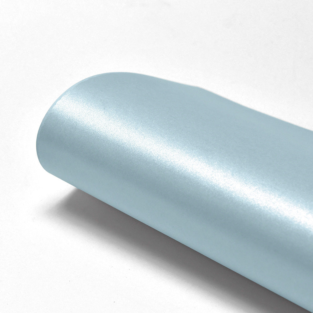 Ein aufgerollter Bogen ☆ hellblaues Schimmer-Effekt Papier (310g/m²), ideal als Papier für Karten, liegt auf einer weißen Fläche und zeigt seine metallisch glänzende Oberfläche.