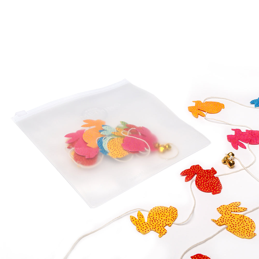 Eine durchsichtige Reißverschlusstasche von ☆ mit farbenfrohen Ausschnitten in Hasenform, die neben einer handgefertigten Girlande aus nepalesischem Papier gezeigt wird.