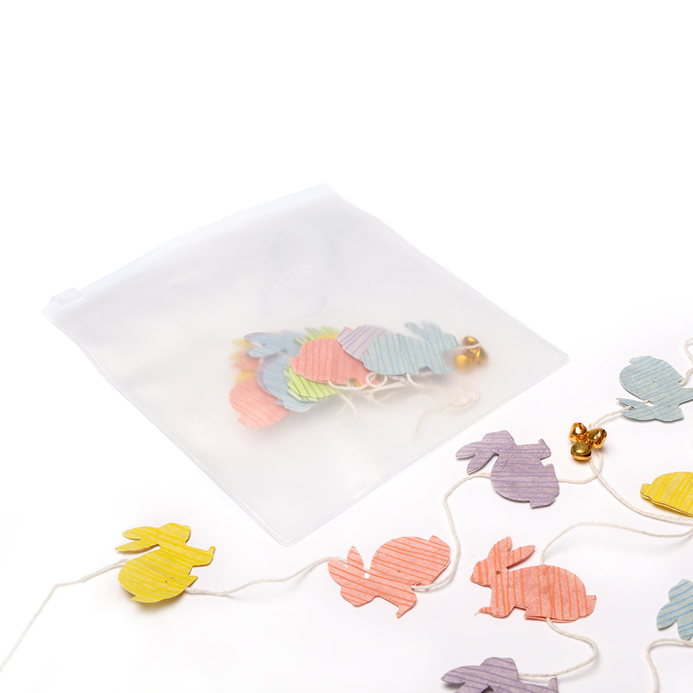 Pastellfarbene Hasenschnurgirlande mit Glöckchen von ☆, hergestellt aus nachhaltigem und handgeschöpftem Papier, teilweise in einem durchsichtigen Reißverschlussbeutel auf weißem Hintergrund.