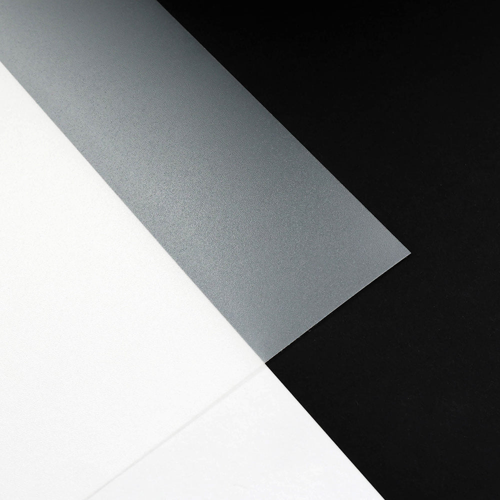 Drei Blätter von Alexandra Renke - weiß, grau-transluzent und schwarz - überlappen sich auf einer Fläche und erinnern an Materialien für die Gestaltung von Buchdeckeln oder Schutzfolien in Buchbindearbeiten.