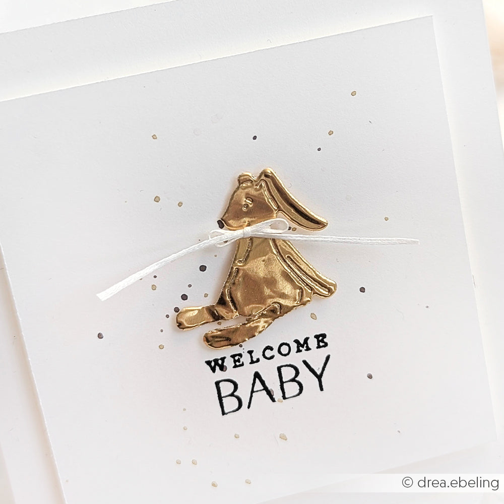 Alexandra Renke Goldhase mit weißer Schleife und "Welcome Baby"-Text auf weißer Karte, mit dezenten goldenen Akzenten und zarten Sprenkeln - ideal für Bastelarbeiten oder handgemachte Geschenke.