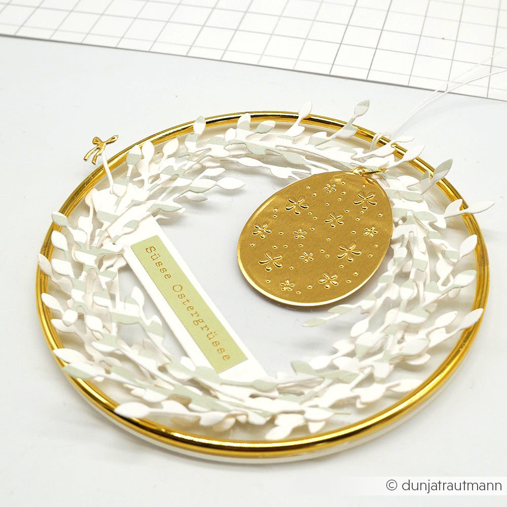 Goldring mit weißen Papierblättern, ein goldener Eierschmuck mit Prägefolie Aluminium für goldene Akzente und ein Stille Ostergrüße"-Anhänger - perfekt für kreative Bastelarbeiten von Alexandra Renke.