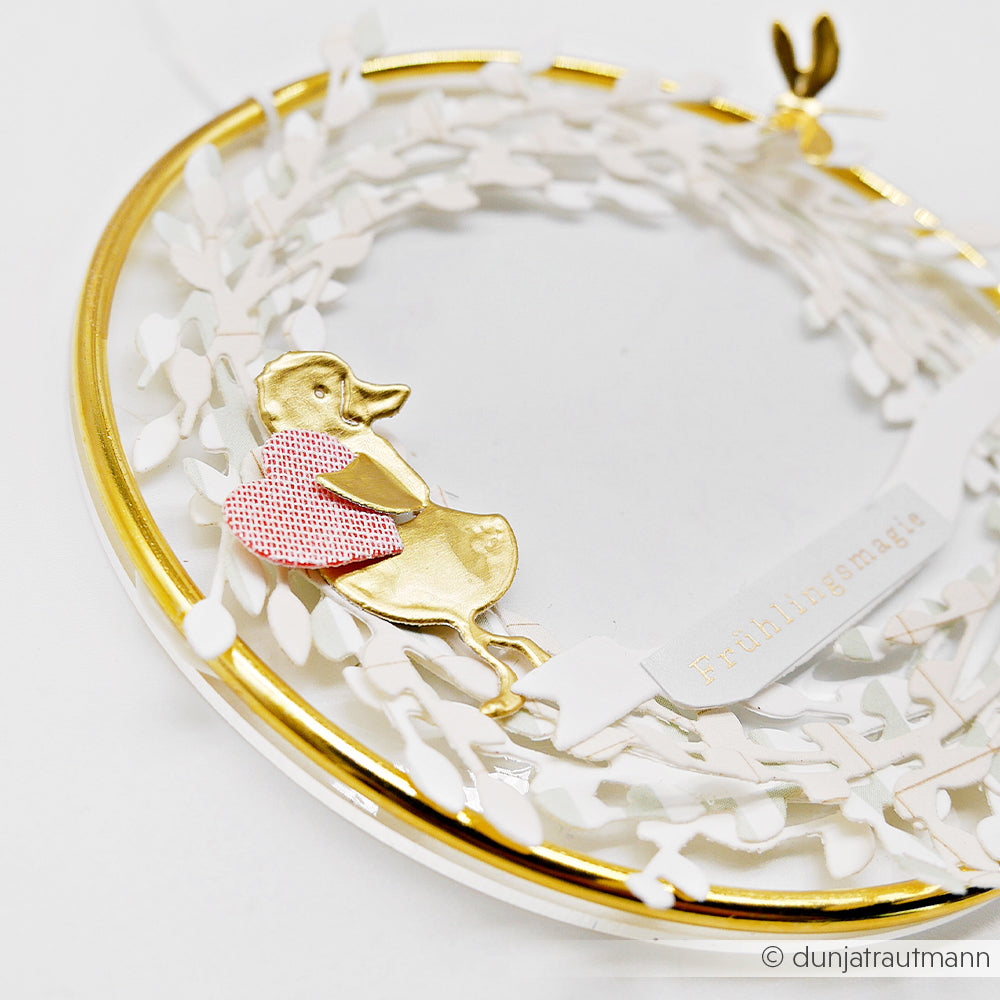 Die goldene Ente mit rosa Herz von Alexandra Renke sitzt auf einem weiß-goldenen Kranz mit Goldfolien-Akzenten und einem "Frühlingsmarie"-Banner, perfekt für besondere Bastelprojekte.