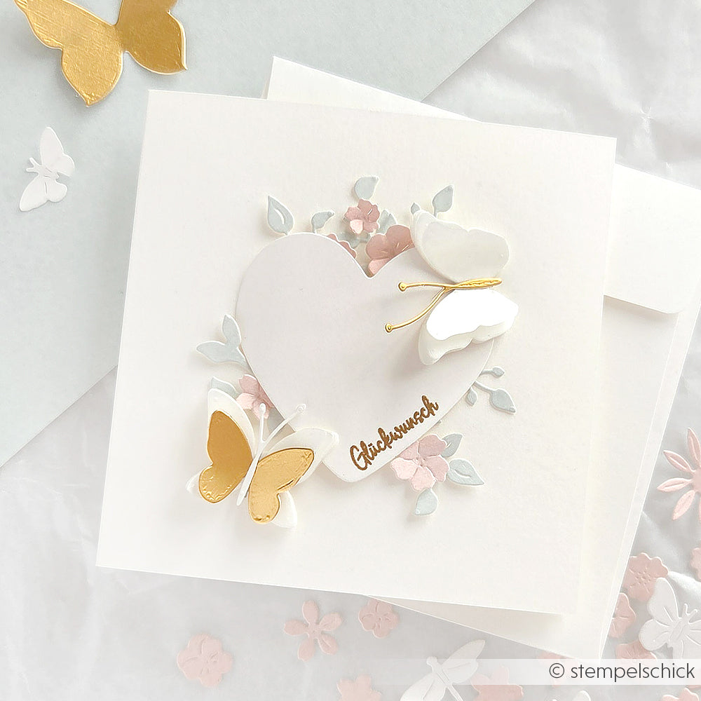 Die quadratische Karte von Alexandra Renke zeigt ein weißes Herz mit dem Text "Glückwunsch", pastellfarbene Blumen sowie goldene und weiße Schmetterlinge. Goldene Akzente und zarte Handarbeit verleihen der Karte einen besonderen Charme.