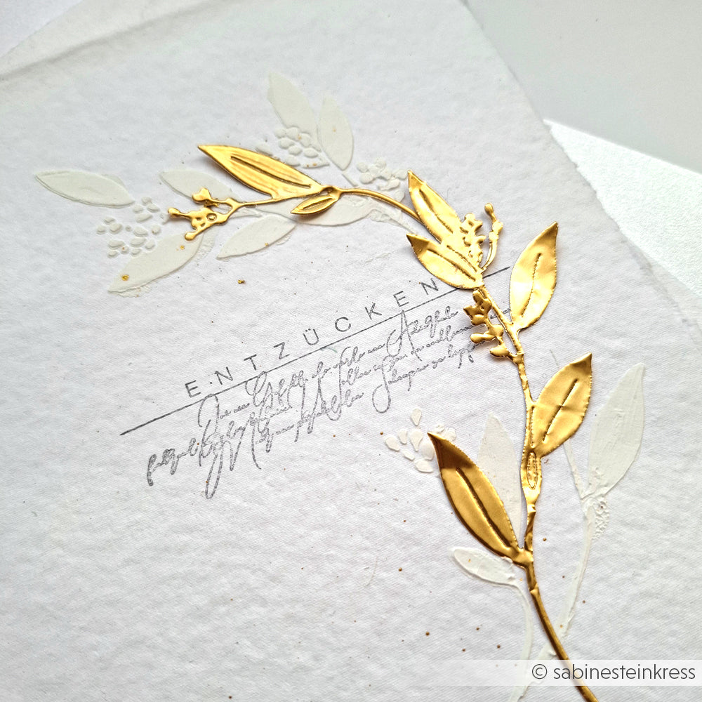 Handgefertigte Alexandra Renke Karte mit gold-weiß geprägten Blättern, Zierschrift und "ENTZÜCKEN" in der Mitte - ideal für Bastelarbeiten-Fans, die goldene Akzente lieben.