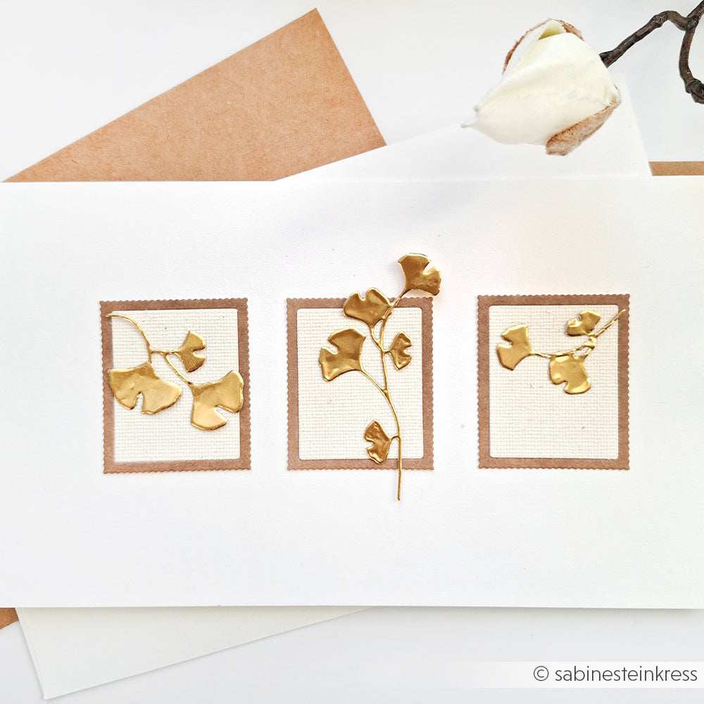Eine handgefertigte Karte von Alexandra Renke mit drei goldenen Ginkgoblättern aus Prägefolie Aluminium, gerahmt auf weißem und braunem Papier mit goldenen Akzenten, wird neben einem Umschlag und einer Blume präsentiert.