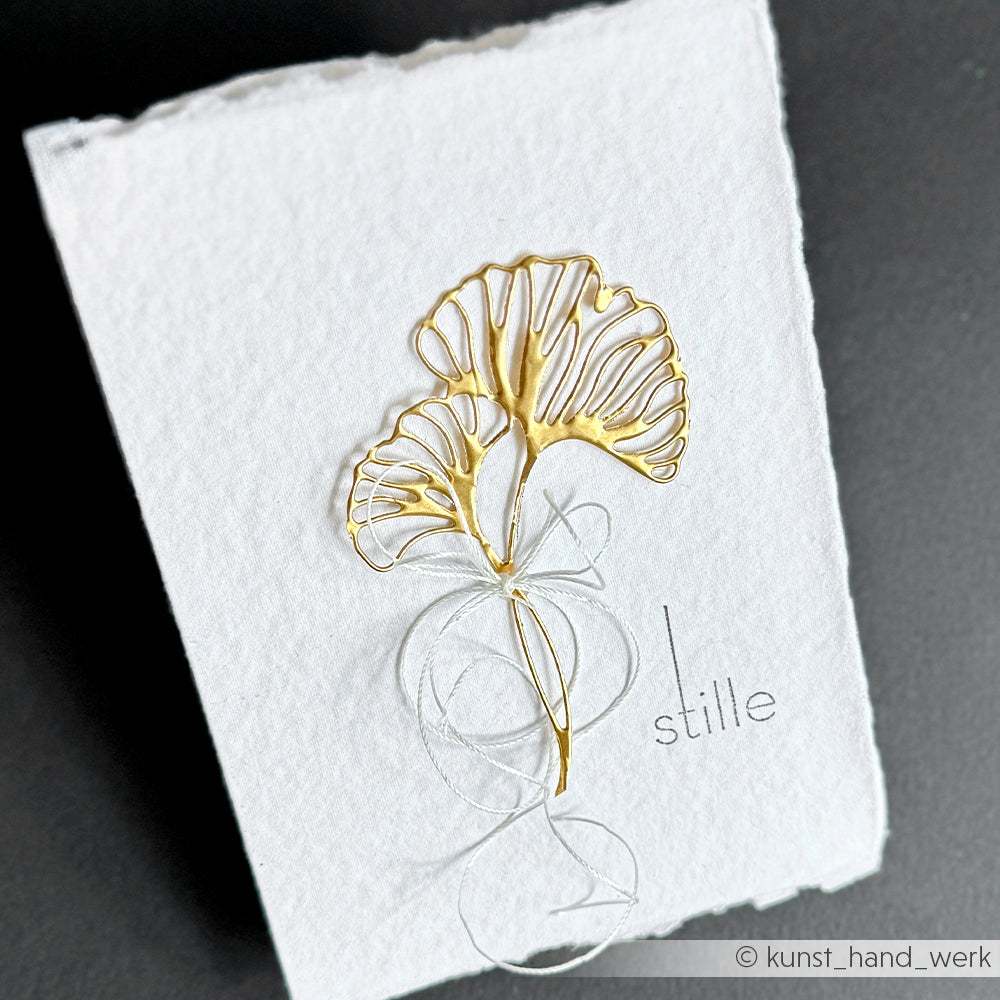 Zarte Gingko-Blätter aus Golddraht mit goldenen Akzenten auf strukturiertem weißem Papier von Alexandra Renke, mit dem Wort "stille" neben dem Design gedruckt.