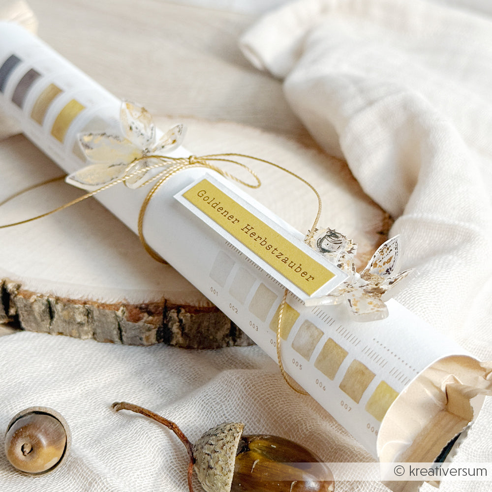 Ein in weißes Papier mit goldenen Akzenten und Herbstblättern eingewickeltes Geschenk, das auf rustikalem Holz liegt – eine perfekte Idee, um handgefertigte Schätze wie selbstgemachten Schmuck oder selbstgebastelte Schlüsselanhänger zu präsentieren, die vielleicht aus bunter Schrumpffolie gefertigt wurden.