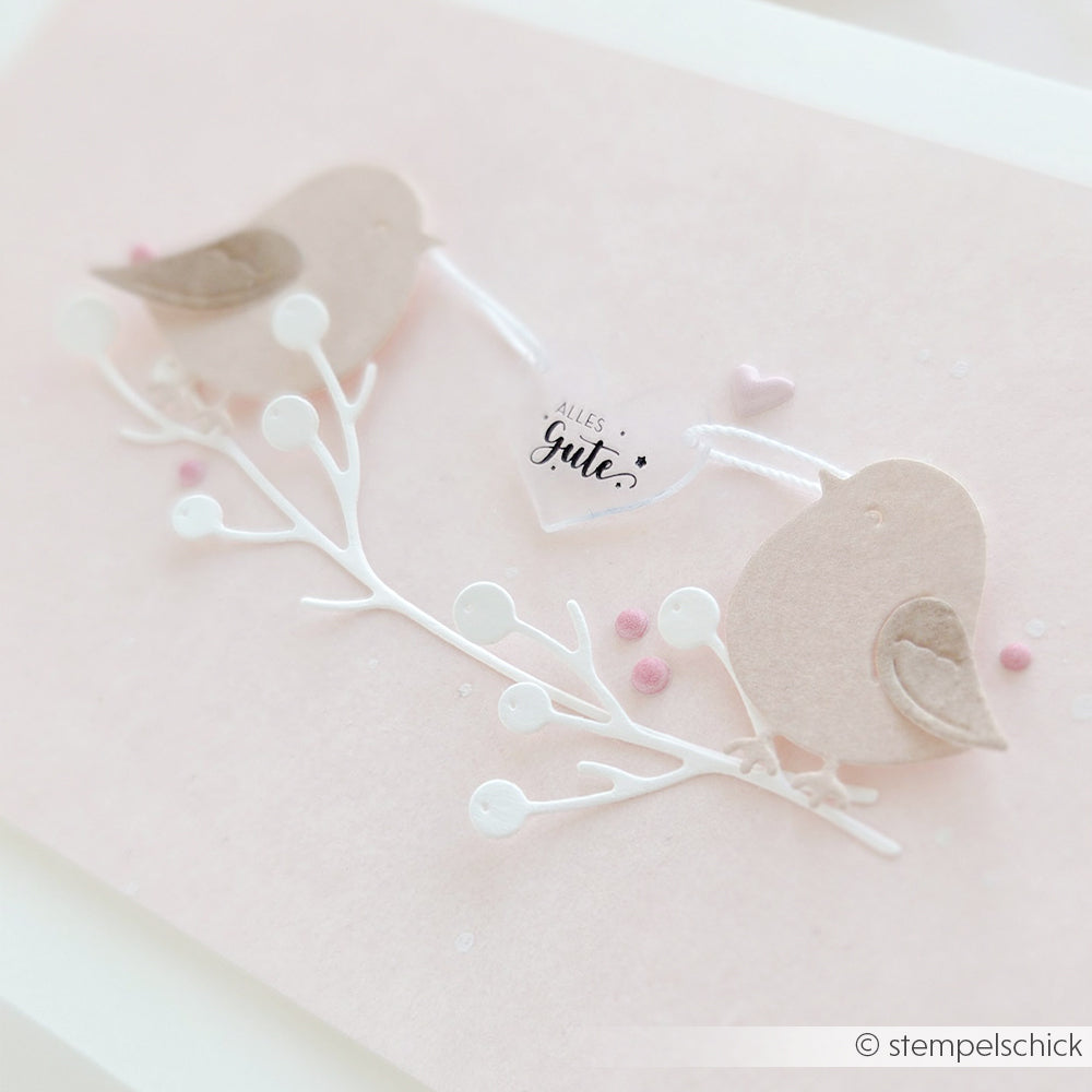 Zwei Papiervögel auf einem weißen Ast mit einem Anhänger, auf dem „Alles Gute“ steht, vor einem pastellrosa Hintergrund – die perfekte Inspiration, wenn Sie Schmuck basteln oder Anhänger selbst gestalten möchten, vielleicht sogar mit Schrumpffolie für kreative Effekte.
