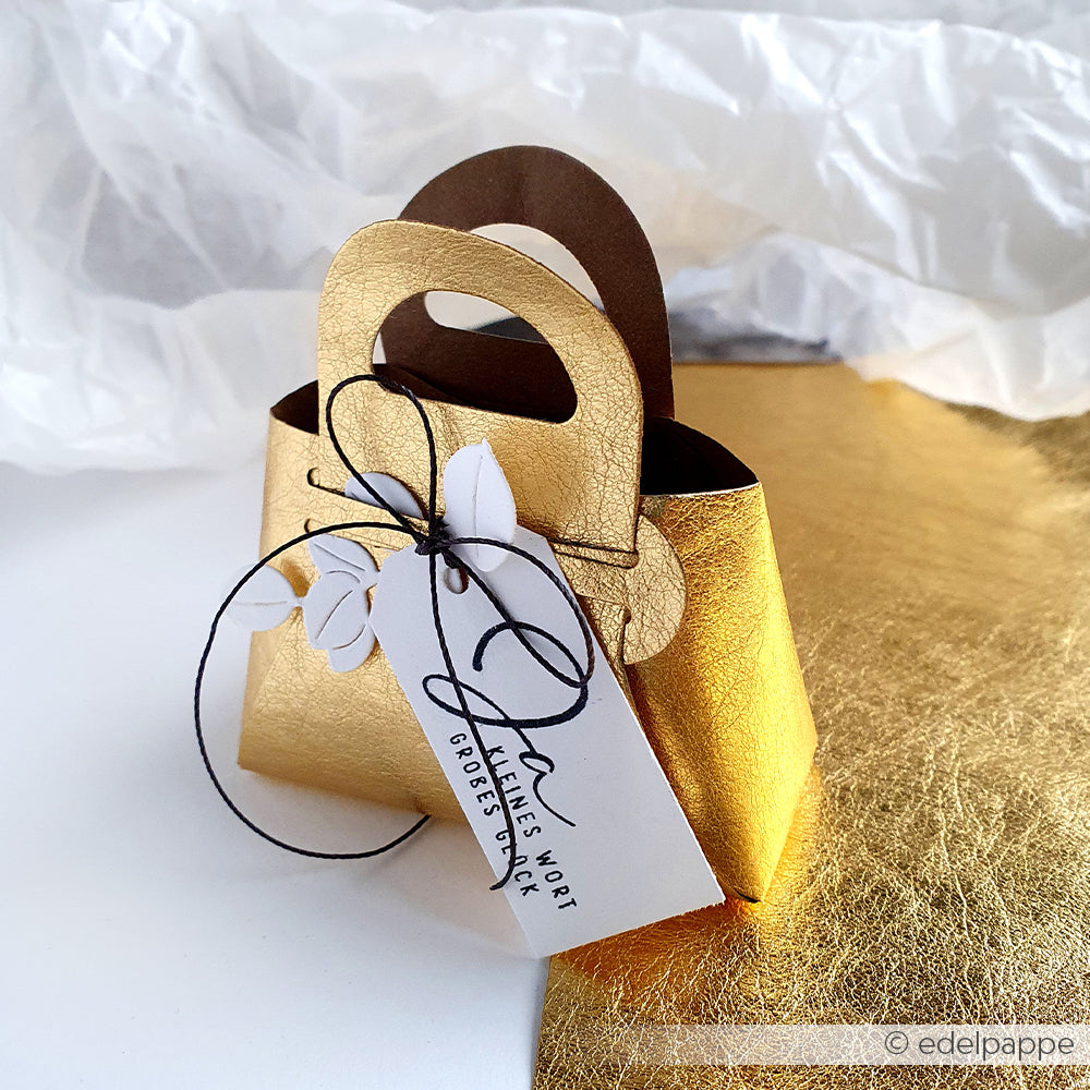 Eine kleine, goldene, taschenförmige Geschenkbox von Alexandra Renke aus seidenmatt glänzendem Papier mit einem handgeschriebenen Anhänger und schwarzer Kordel sitzt auf einer goldenen Oberfläche.