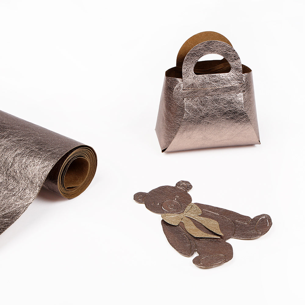 Metallisch strukturiertes PAPP-Papier, eine kleine Geschenktüte und ein flacher Teddybär-Ausschnitt von Alexandra Renke auf weißem Untergrund - perfektes Buchbinder Zubehör für kreative Projekte.