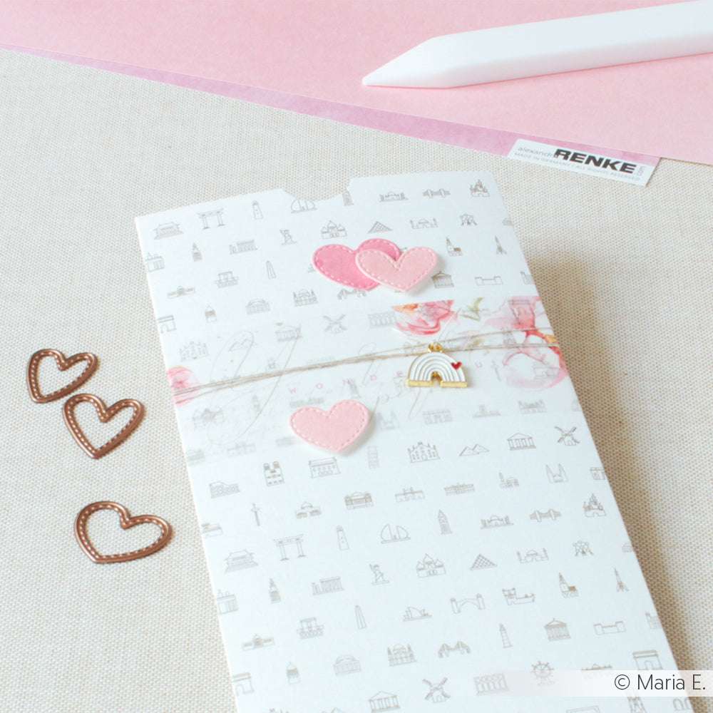 Dekorativer Umschlag mit rosa Herzverzierungen, Herz-Büroklammern, Buchbinde-Zubehör und einem weißen Werkzeug von Alexandra Renke auf einem pastellfarbenen Schreibtisch.