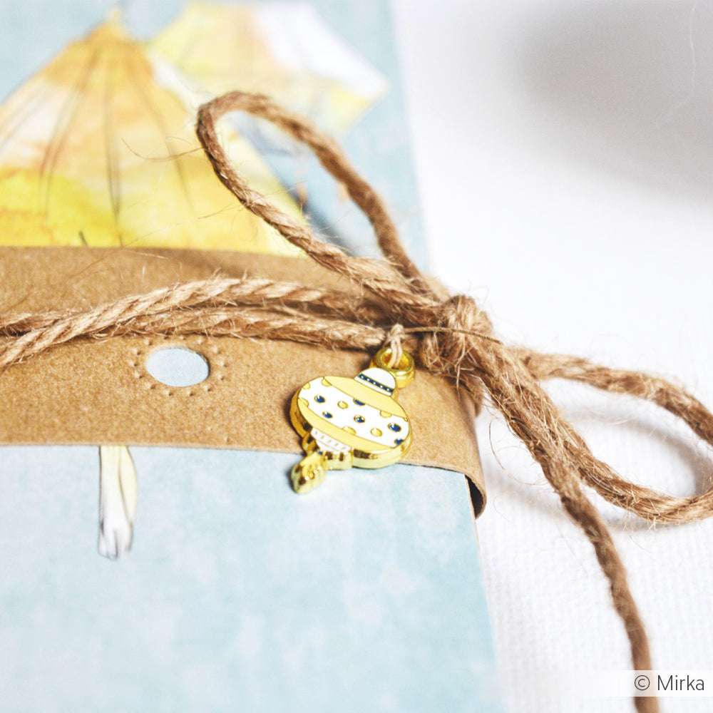 Eine braune Kordelschleife mit einem gelben Lampion-Charm, der um eine blaue Karte mit einer gelben Figur gebunden ist - dieses Accessoire von Alexandra Renke ist eine reizvolle Ergänzung für Buchbindearbeiten oder kreative Geschenkanhänger.