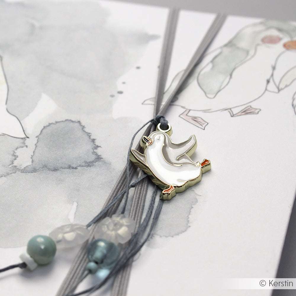 Nahaufnahme eines weißen Vogelanhängers von Alexandra Renke an einer mehrreihigen Halskette, die auf einer illustrierten Karte mit Vogelmotiv abgebildet ist.