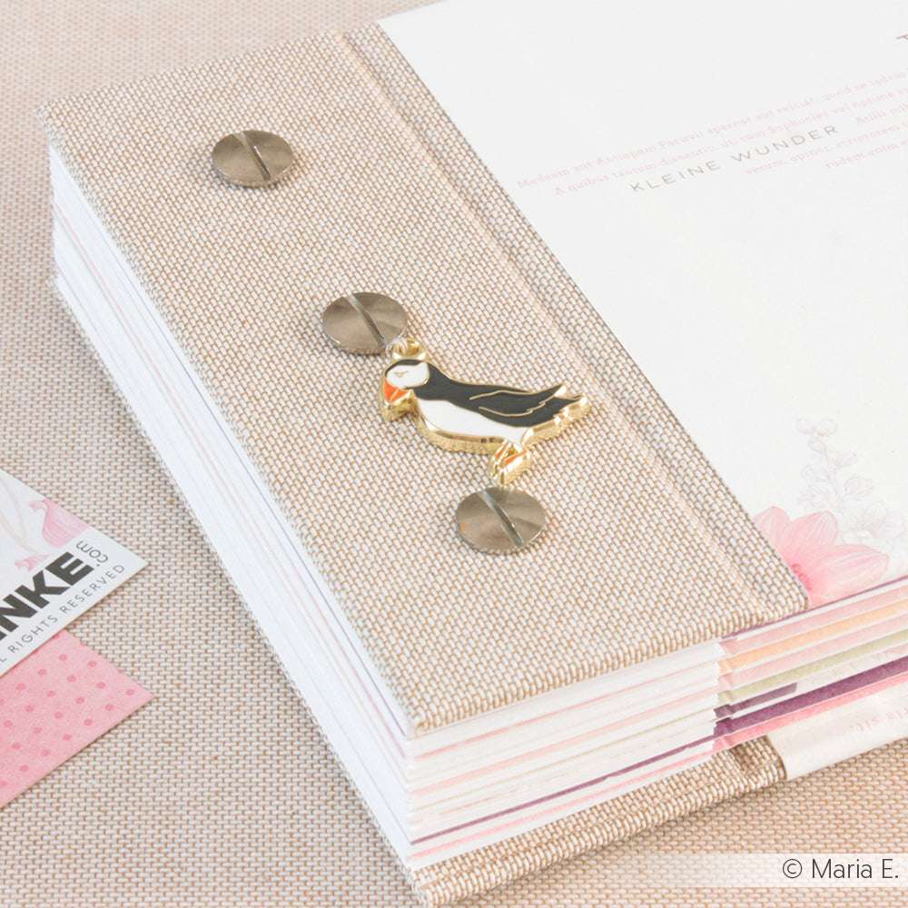 Ein beigefarbenes stoffgebundenes Notizbuch von Alexandra Renke, das mit der Technik des Buchbindens hergestellt wurde, ist mit einer goldenen Charm Puffin-Anstecknadel und Metallnieten auf dem Einband versehen und liegt teilweise geöffnet auf einem Schreibtisch.