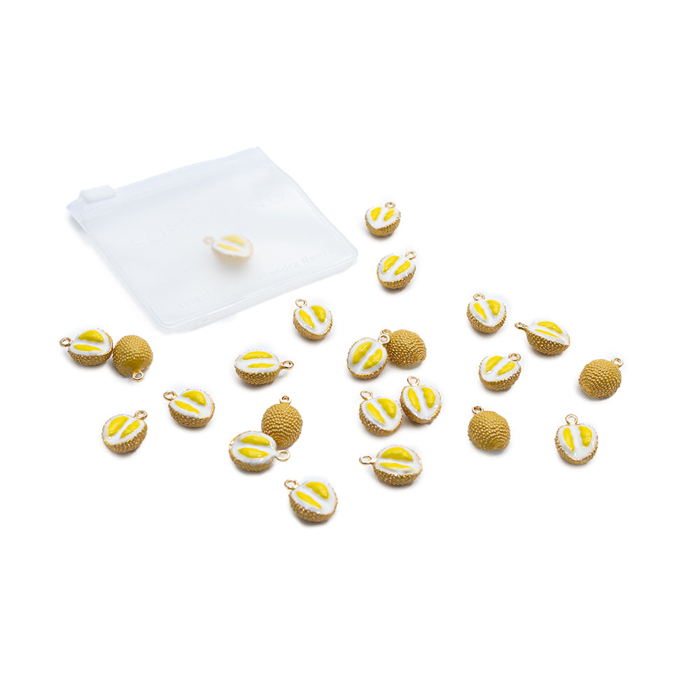 Neben einem durchsichtigen Plastikbeutel liegen kleine Obst-Anhänger mit Zitronenmotiv von Alexandra Renke sowie strukturierte Goldschmuckstücke verstreut.