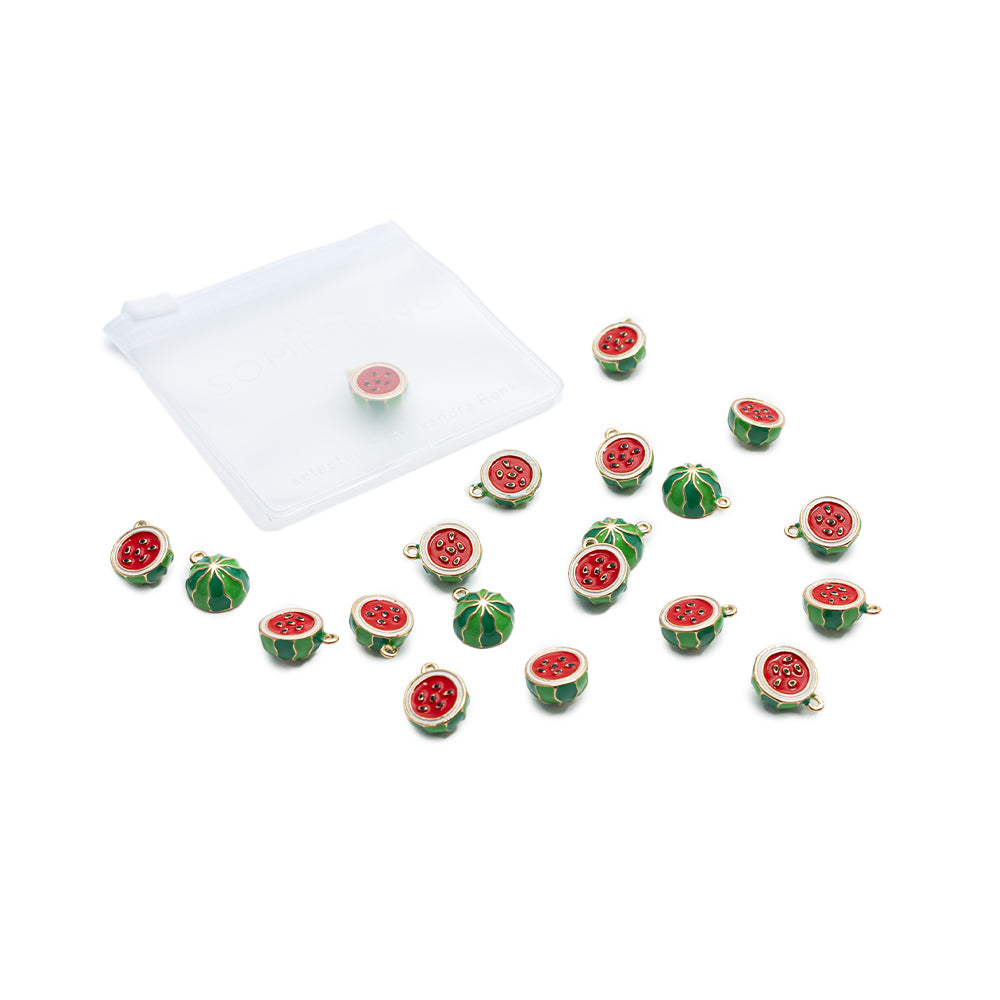 Kleine, wassermelonenförmige „Obst“-Charms und Anhänger von Alexandra Renke, verstreut auf einer weißen Fläche, im Hintergrund ist ein durchsichtiges Reißverschluss-Säckchen zu sehen.