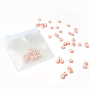 Kleine rosafarbene und weiße Perlen zum Auffädeln von Alexandra Renke sind in der Nähe eines durchsichtigen Reißverschlussbeutels auf weißem Hintergrund verstreut, ideal um Schmuck selbst zu machen.