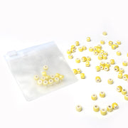 Kleine gelbe und weiße zylindrische Perlen von Alexandra Renke, perfekt für die Schmuckherstellung oder zum Basteln, verstreut in einem durchsichtigen Beutel mit Reißverschluss auf weißem Hintergrund.