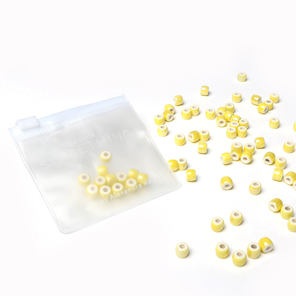 Kleine gelbe und weiße zylindrische Perlen von Alexandra Renke, perfekt für die Schmuckherstellung oder zum Basteln, verstreut in einem durchsichtigen Beutel mit Reißverschluss auf weißem Hintergrund.