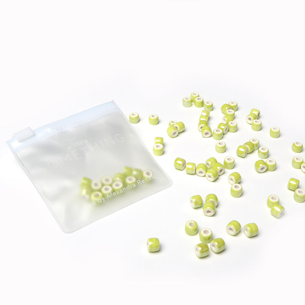 Kleine grün-weiß glasierte Keramikperlen von Alexandra Renke sind in einem transparenten, wiederverschließbaren Beutel auf weißem Hintergrund verstreut - ideal für Perlen basteln oder Schmuck selber machen.