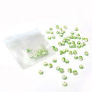 Kleine grüne und weiße Alexandra Renke Perlen basteln verstreut neben einem halbtransparenten Reißverschlussbeutel auf weißem Hintergrund - ideal für Schmuck selbst machen Projekte.