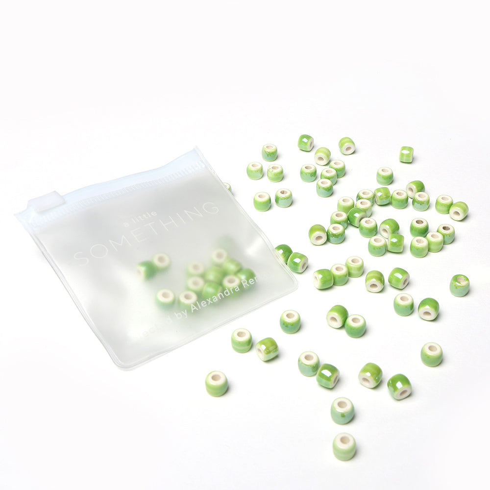 Kleine grüne und weiße Alexandra Renke Perlen basteln verstreut neben einem halbtransparenten Reißverschlussbeutel auf weißem Hintergrund - ideal für Schmuck selbst machen Projekte.