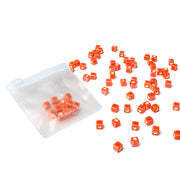 Kleine orangefarbene Würfelperlen von Alexandra Renke, verstreut auf einer weißen Oberfläche, einige in einem durchsichtigen Beutel - ideal als glasierte Keramikperlen für die Schmuckherstellung oder als Fädelperlen für kreative Projekte.