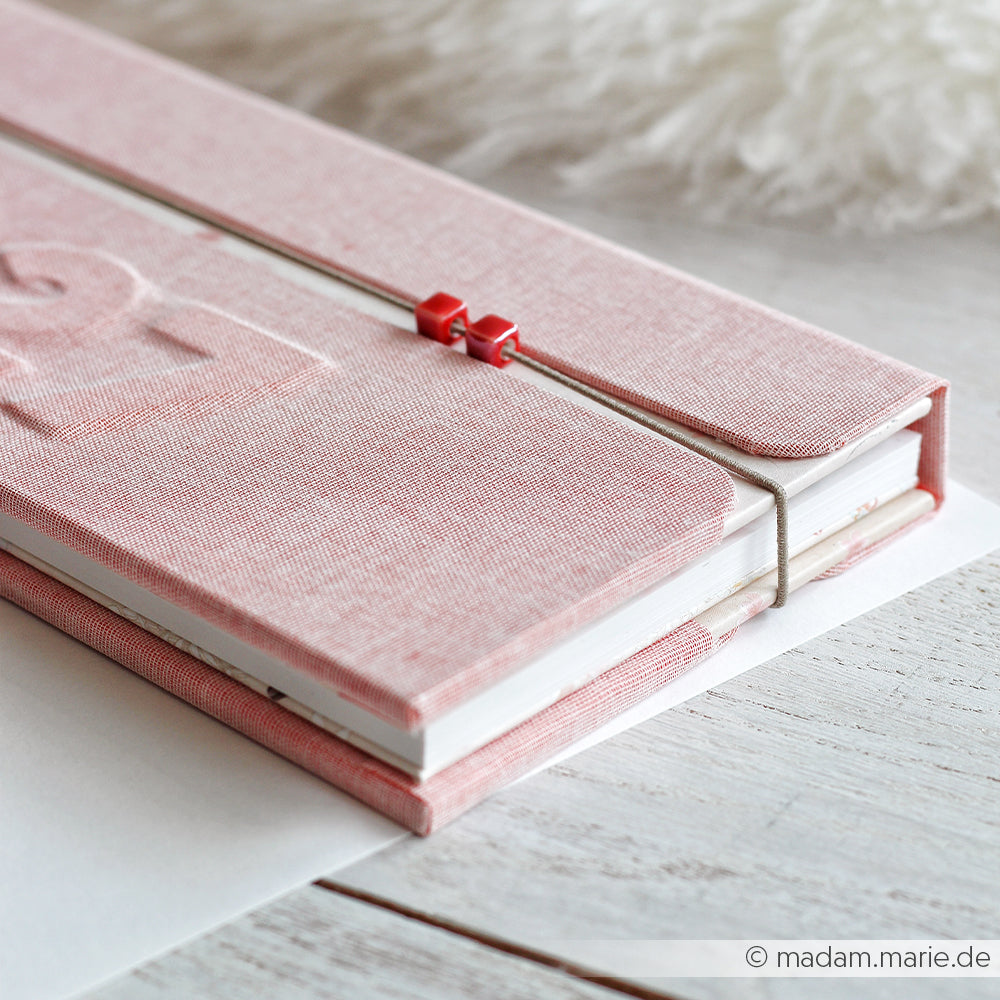Ein mit rosa Stoff bezogenes Notizbuch mit Gummiband und rotem Verschluss, das auf einer weißen Unterlage liegt – ideal, um kreative Ideen für das Perlenbasteln festzuhalten oder dein nächstes „Schmuck selber machen“-Projekt mit glasierten Keramikperlen zu entwerfen.