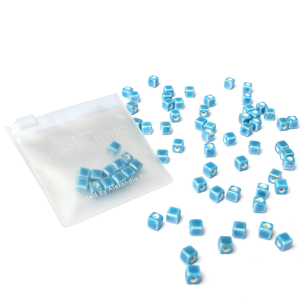 Kleine blau-weiße quadratische Alexandra Renke Perlen zum Auffädeln in der Nähe eines durchsichtigen Reißverschlussbeutels auf weißem Hintergrund - perfekt für Schmuck selber machen.