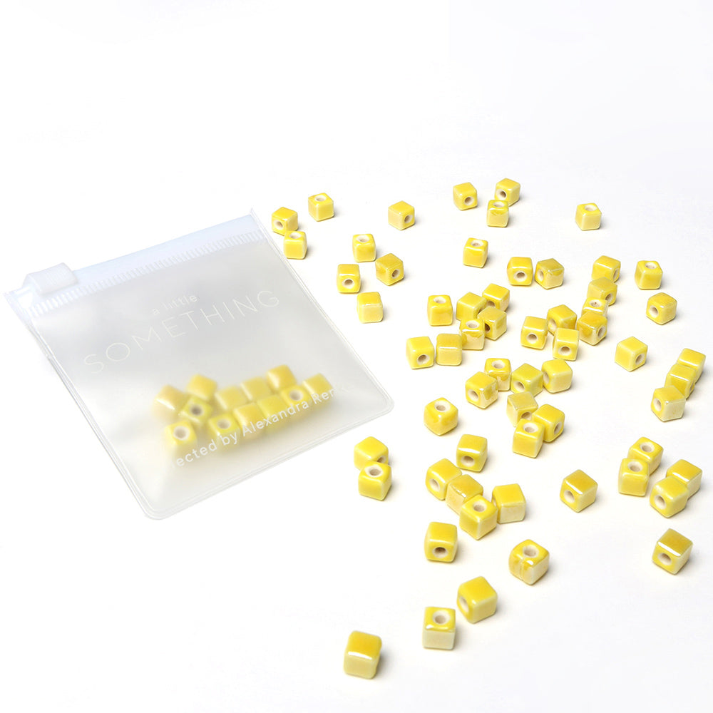 Kleine gelbe Keramikperlen-Würfel von Alexandra Renke verstreut neben einem durchsichtigen Reißverschlussbeutel auf weißem Hintergrund - ideal für glasierte Schmuckgestaltungsprojekte.