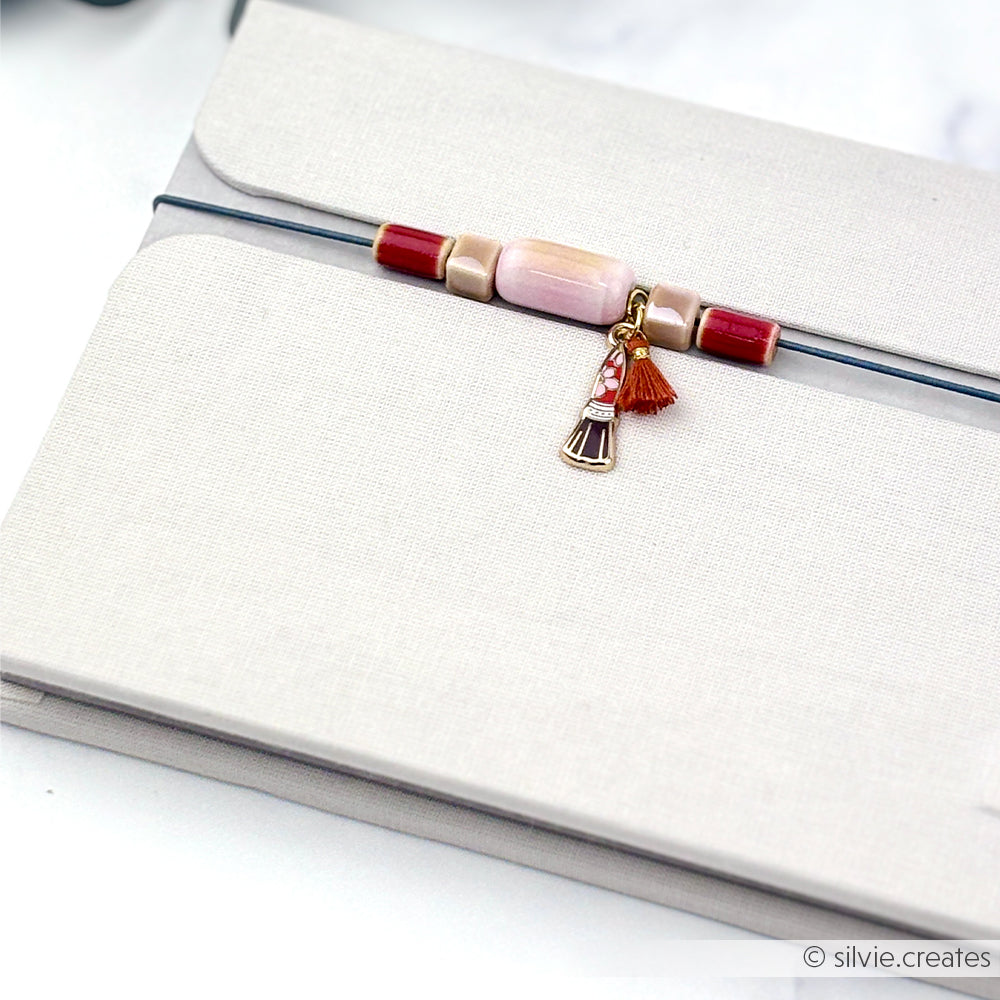 Ein beiges Notizbuch mit einem Anhänger aus glasierten Keramikperlen, der mit Perlen und Quasten verziert ist und an einem elastischen Band befestigt ist, auf einem weißen Hintergrund – ein perfektes Accessoire für alle, die gerne selbst Schmuck basteln oder nach einzigartigen Perlen zum Auffädeln suchen.