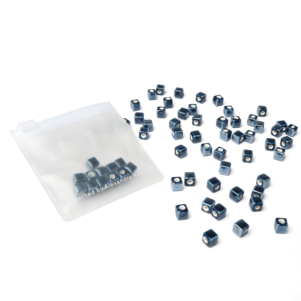 Kleine blaue glasierte Keramikperlen von Alexandra Renke verstreut neben einem halbtransparenten Kunststoffbeutel auf weißem Hintergrund, perfekt zur Schmuckherstellung.