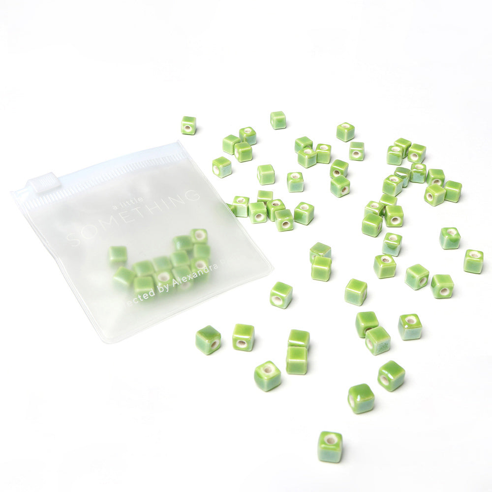 Die kleinen grünen Würfelperlen von Alexandra Renke, die sich perfekt zum Perlenbasteln und zur Schmuckgestaltung eignen, werden neben einem durchsichtigen, wiederverschließbaren Beutel auf weißem Hintergrund präsentiert.