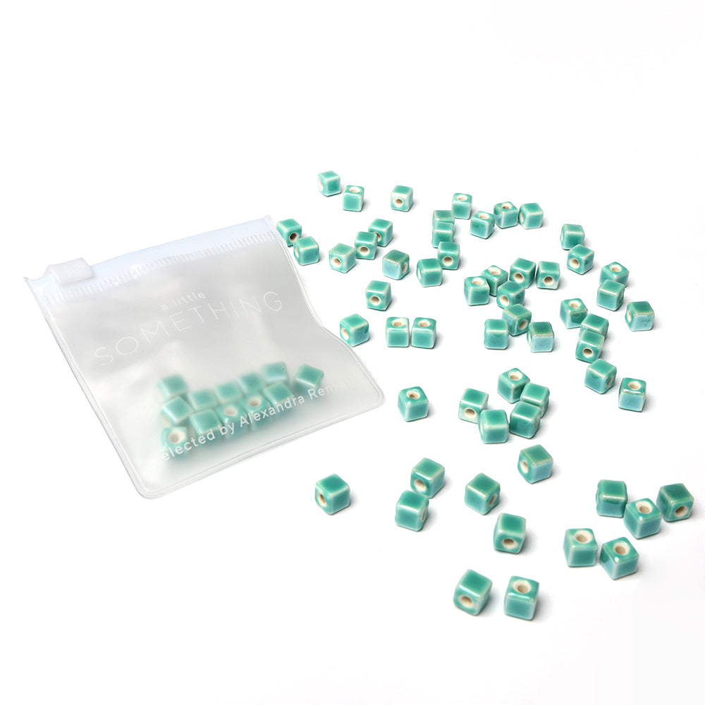 Kleine tealfarbene, glasierte Keramikwürfel von Alexandra Renke tropfen aus einem durchsichtigen Beutel auf eine weiße Oberfläche - ideal für die Schmuckherstellung oder das Basteln mit Keramikperlen.