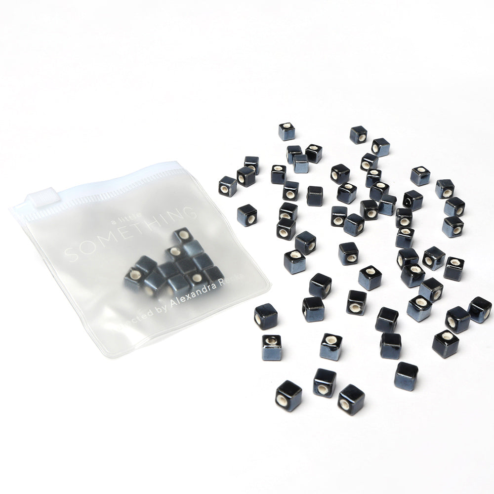 Kleine schwarze, glasierte würfelförmige Keramikperlen mit weißen Punkten von Alexandra Renke, teils verstreut und teils in einem durchsichtigen Zip-Beutel auf einer weißen Fläche - ideal für Schmuck selber machen.