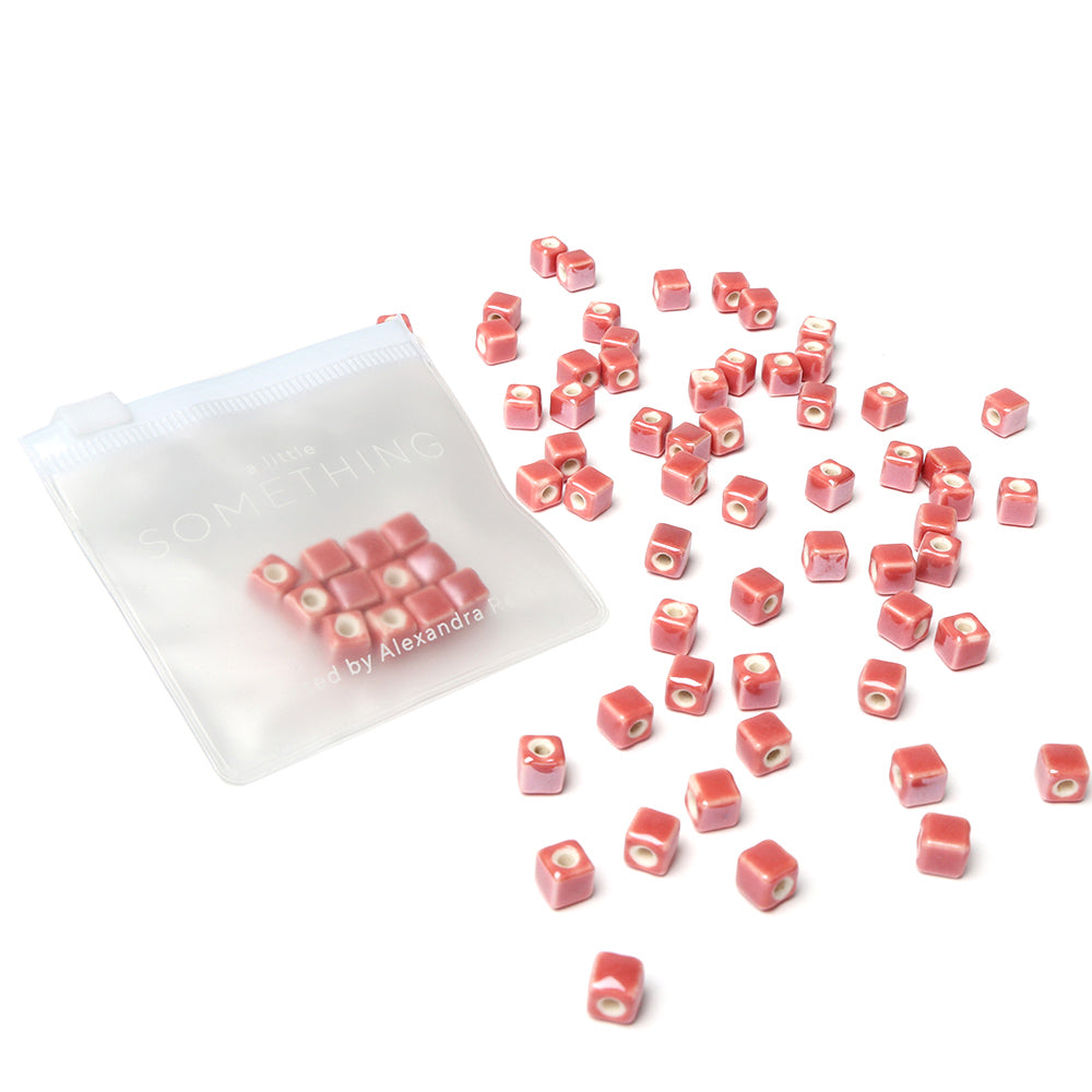 Kleine rosa Würfelperlen von Alexandra Renke, ideal zum Perlen basteln, sind neben einem transparenten Reißverschlussbeutel auf weißem Hintergrund verstreut. Perfekt für alle, die gerne Schmuck selbst machen.