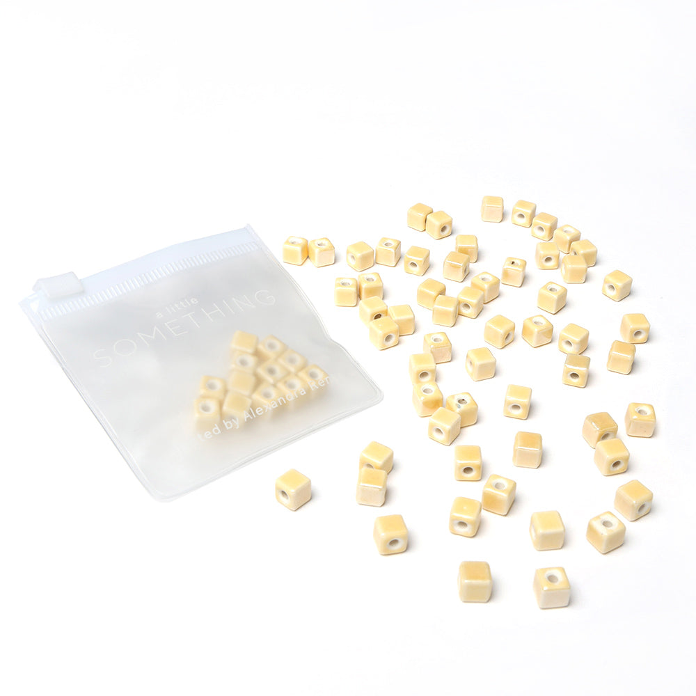 Kleine beige glasierte Keramikperlen von Alexandra Renke verstreut neben einem teilweise offenen durchsichtigen Reißverschlussbeutel auf Weiß - ideal für Schmuckherstellung oder Perlen basteln Projekte.