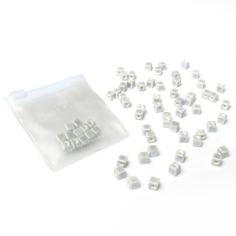 Kleine weiße quadratische Perlen zum Auffädeln von Alexandra Renke liegen neben einem transparenten Zip-Beutel mit der Aufschrift "A Little Something" - ideal, um eigenen Schmuck zu gestalten.