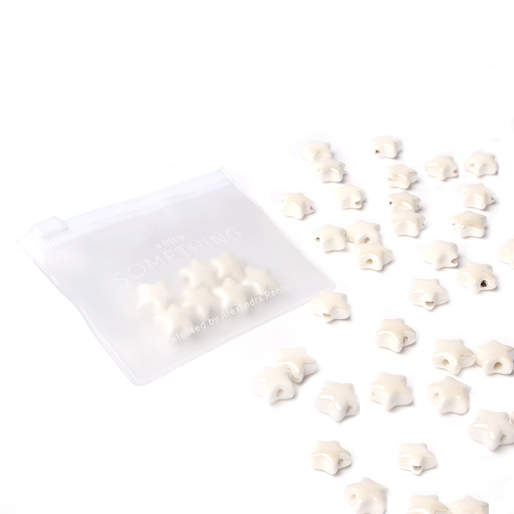 Kleine weiße Sternform-Perlen von Alexandra Renke, die wie glasierte Keramik aussehen, sind neben einem durchsichtigen Ziplock-Beutel auf weißem Hintergrund angeordnet - perfekt, wenn Sie Schmuck selbst machen möchten.